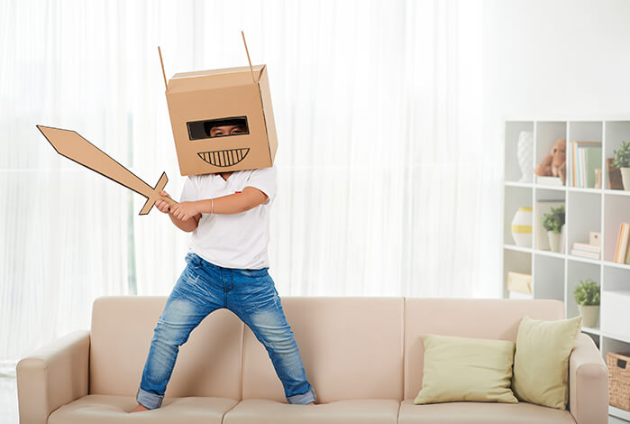 Boy playing with cardboard sword and headgear standing on light brown couch with white curtains in background as well as white bookcase